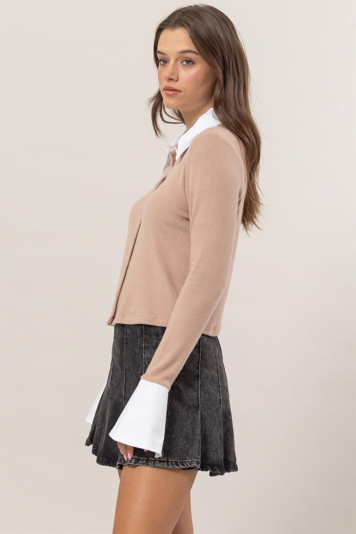 A person with long brown hair is wearing the stylish HYFVE Collared Contrast Button Down Crop Knit Cardigan, a versatile piece featuring a beige design with white cuffs over a collared button-down shirt. They have paired it with a dark grey skirt and are standing sideways against a plain background.