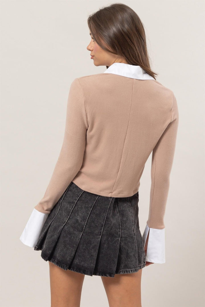 A person wearing a fashionable, adaptable piece—the HYFVE Collared Contrast Button Down Crop Knit Cardigan in beige with white cuffs and collar—paired with a black pleated skirt. They stand against a plain background, facing away.