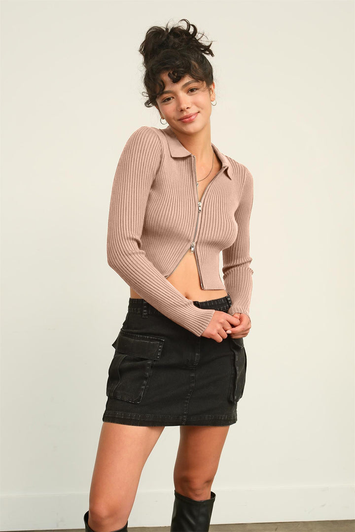 A person wearing the HYFVE Ribbed Double Zip Cropped Cardigan in beige paired with a black denim skirt stands against a light-colored background. Their dark hair is styled in a bun, and they are smiling.