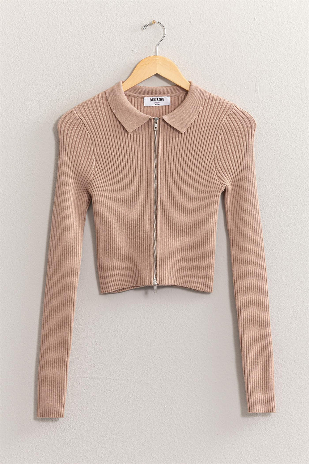 The HYFVE Ribbed Double Zip Cropped Cardigan is an elegant, beige, long-sleeve piece with ribbed fabric and a collar. Displayed on a wooden hanger against a light wall, this versatile cardigan features a front zipper closure and a cropped design.