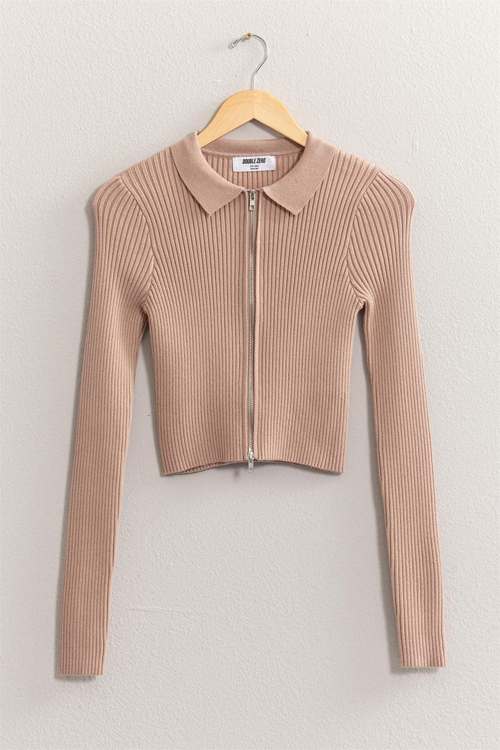 The HYFVE Ribbed Double Zip Cropped Cardigan is an elegant, beige, long-sleeve piece with ribbed fabric and a collar. Displayed on a wooden hanger against a light wall, this versatile cardigan features a front zipper closure and a cropped design.