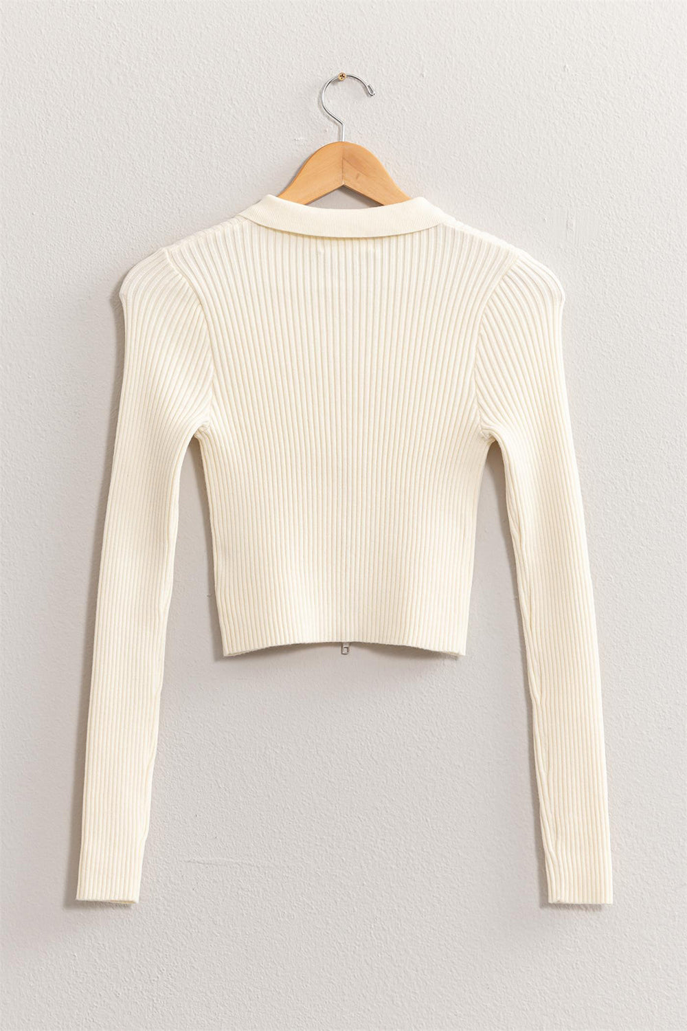 Displayed on a wooden hanger against a light gray wall is the HYFVE Ribbed Double Zip Cropped Cardigan, crafted from ribbed fabric in a white hue. This versatile garment showcases a high neckline and fitted style, perfect for any occasion.