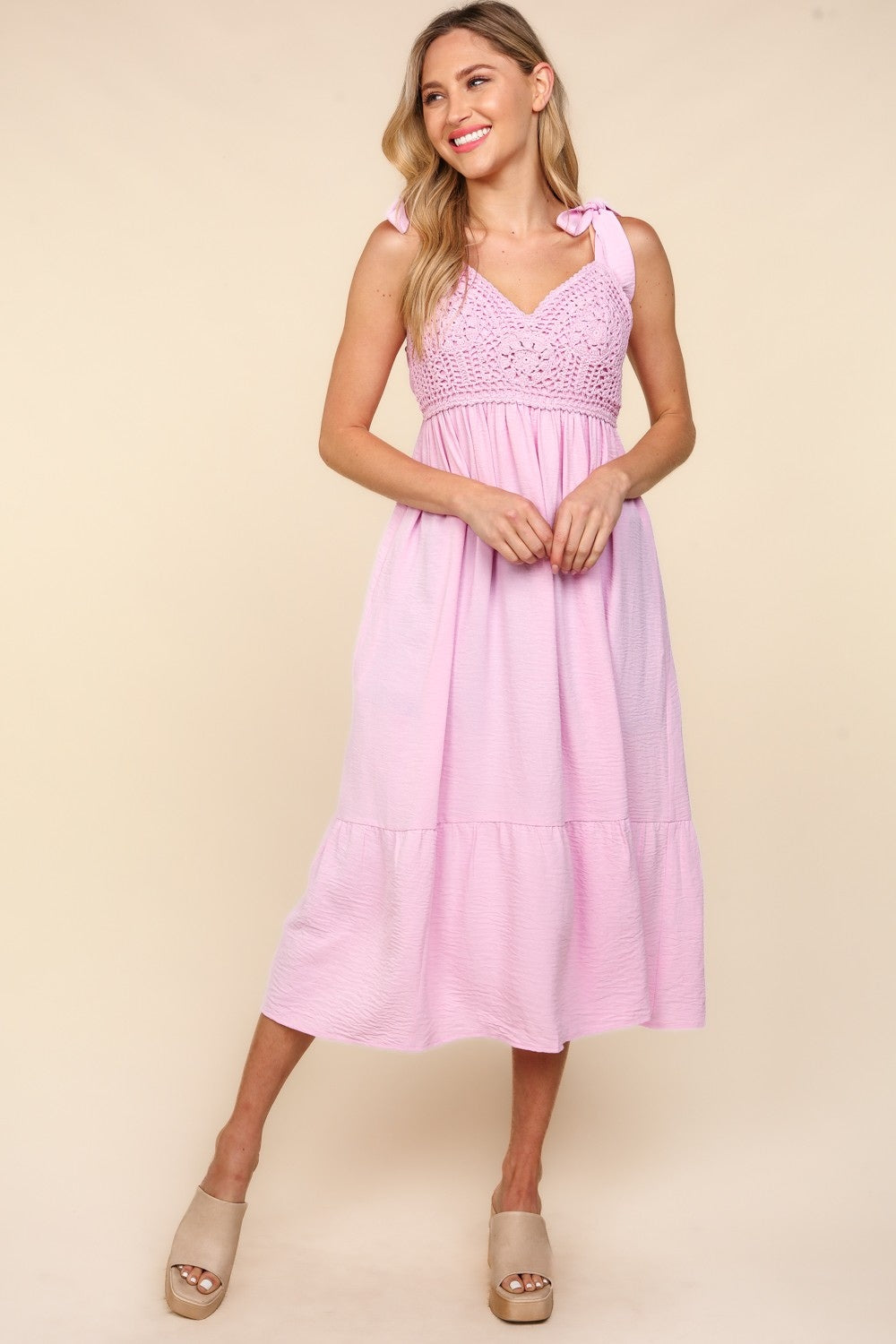 A woman in a pink Haptics Crochet Lace Ruffle Hem Cami Midi Dress with a V-neck is smiling against a neutral background. Her long, light hair cascades as she confidently stands in her wedge sandals.