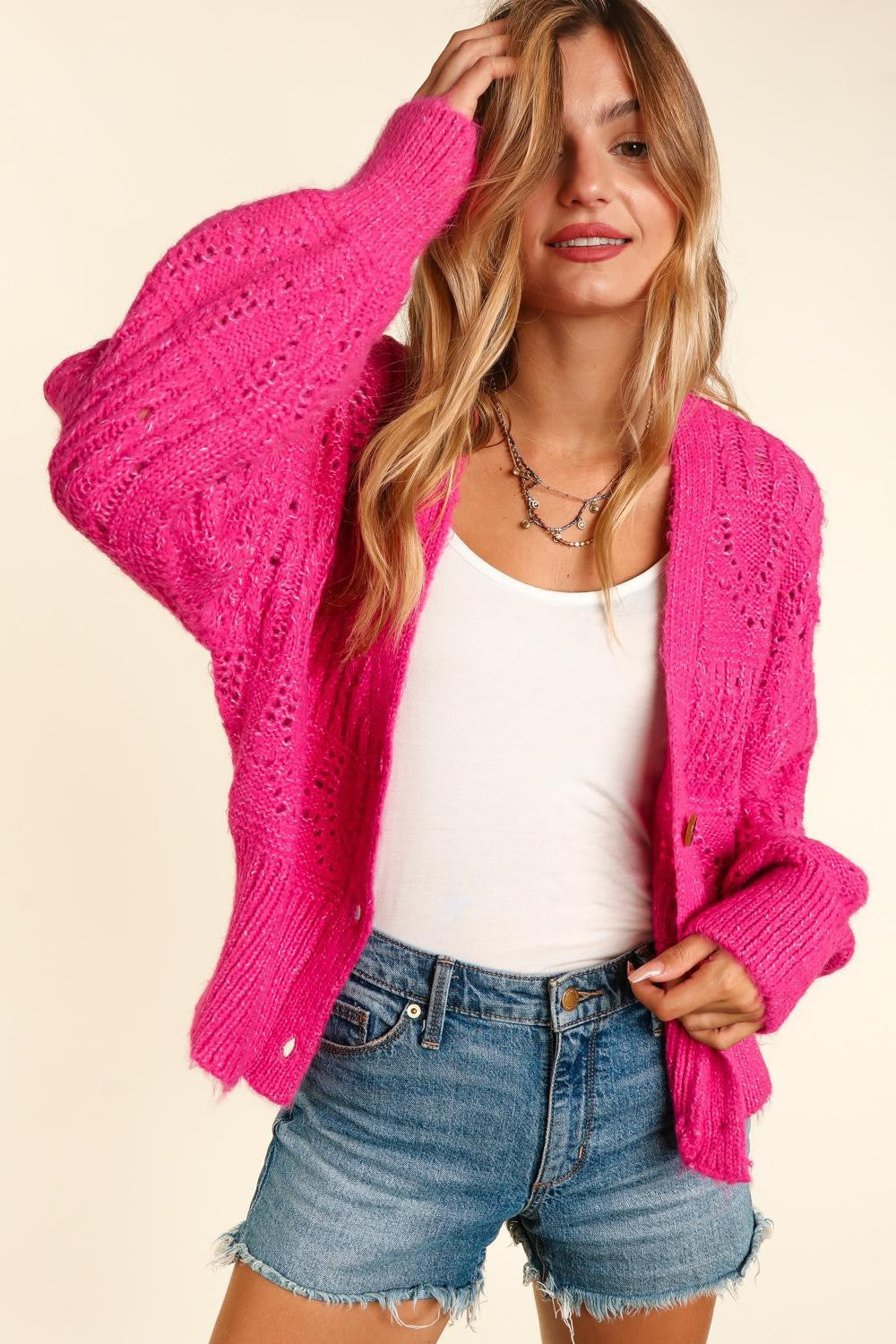 A woman in a chic style showcases a vibrant pink Haptics Full Size Button Down Crop Long Sleeve Cardigan over a white tank top and denim shorts. Her long, wavy hair frames her smiling face as she casually holds her hand to her head against a neutral background.