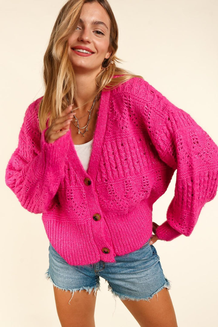 A woman smiles softly, wearing the Haptics Full Size Button Down Crop Long Sleeve Cardigan in chic bright pink paired with denim shorts. The cardigan's loose fit and large brown buttons add an elegant touch to her outfit. She stands against a plain beige background, with her long hair partly covering her face.