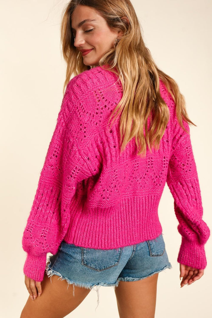 A woman with long hair exudes chic style in a Haptics Full Size Button Down Crop Long Sleeve Cardigan in bright pink, paired with denim shorts. Facing away, she glances over her shoulder with a slight smile against a plain light-colored backdrop.