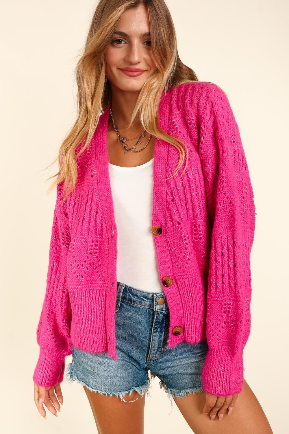 A person wearing a stylish Haptics Full Size Button Down Crop Long Sleeve Cardigan in bright pink over a white top and denim shorts. They have long hair, are smiling, and are standing against a plain background, effortlessly showcasing their chic style.