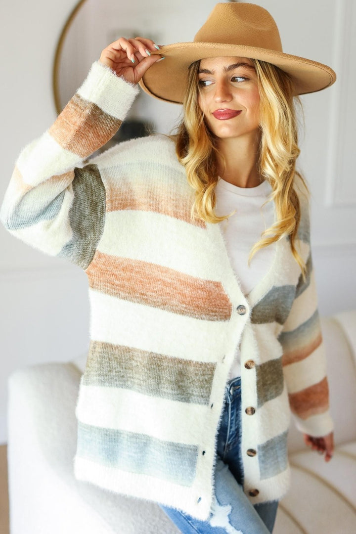 A woman wearing a Haptics Full Size Button Down Stripe Soft Fuzzy Sweater Cardigan and a wide-brimmed tan hat poses with a confident smile, leaning slightly to the side in a bright, cozy room.