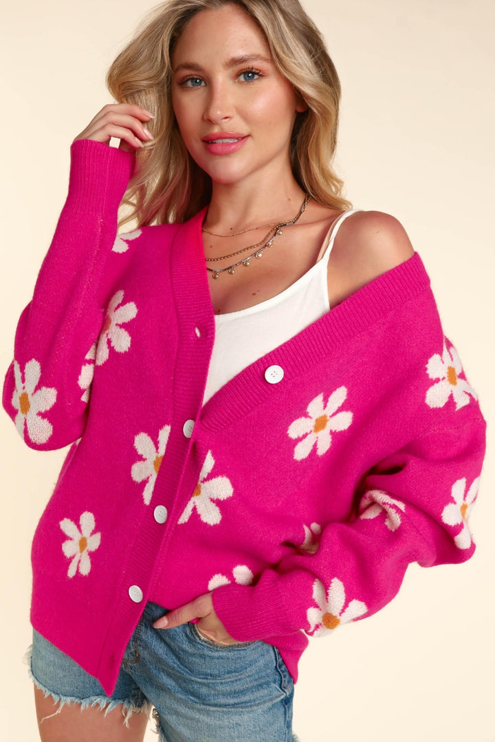 A woman is adorned in the Haptics Full Size Daisy Floral Button Down Long Sleeve Cardigan in a vibrant pink hue, layered over a white tank top. She smiles warmly, with one hand gently touching her collar. Her blonde hair beautifully complements her denim shorts and stacked necklaces against the neutral backdrop.