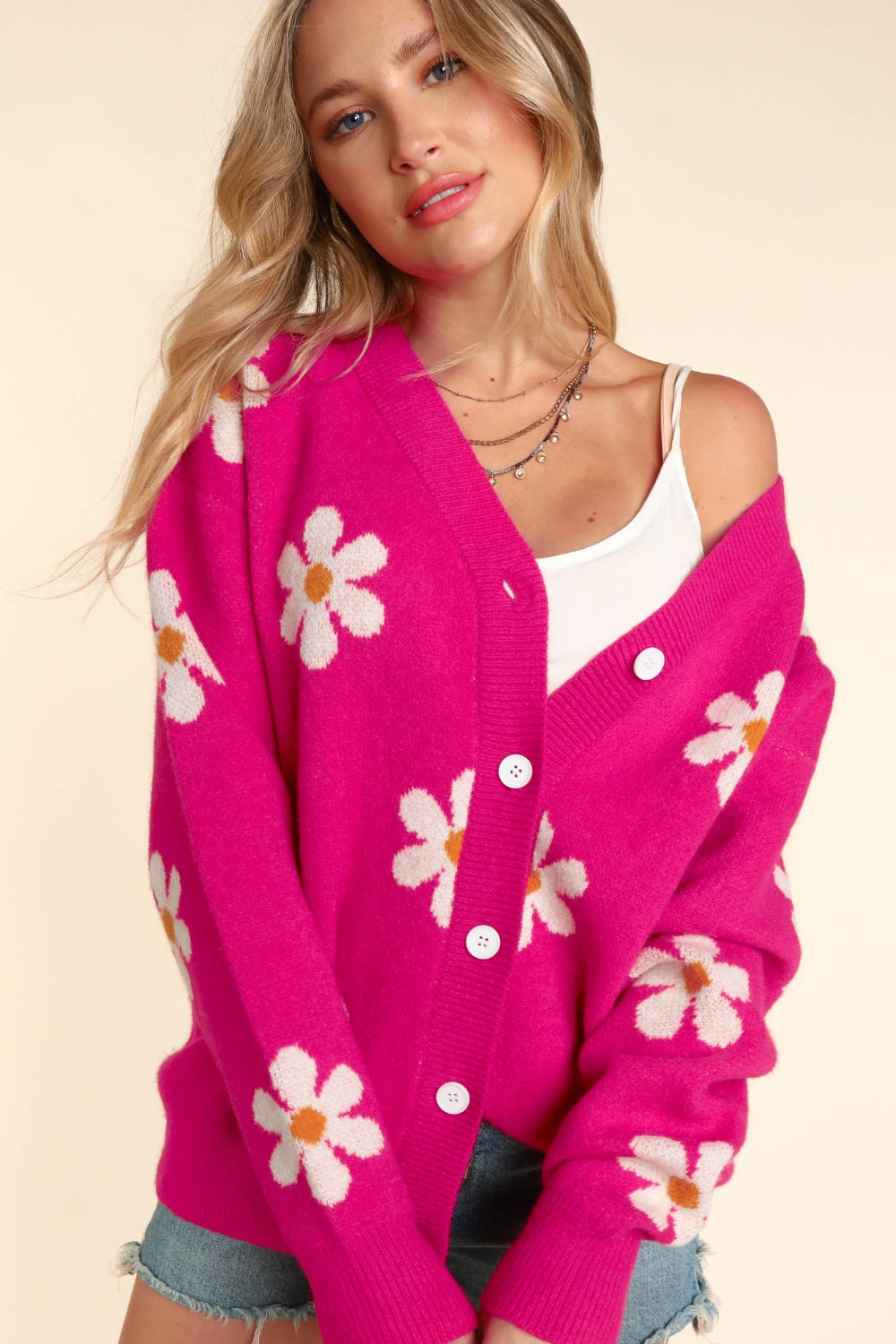 A woman dressed in the Haptics Full Size Daisy Floral Button Down Long Sleeve Cardigan exudes effortless style. The vibrant pink cardigan, adorned with charming white daisy patterns, is slightly slipped off one shoulder to reveal a white tank top. Her long, wavy blonde hair cascades over her layered necklace against a beige background.