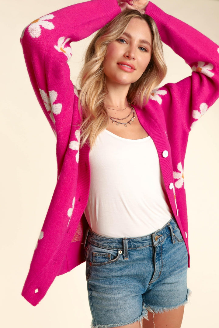 A person confidently poses in a Haptics Full Size Daisy Floral Button Down Long Sleeve Cardigan over a white tank top and denim shorts. Their arms are raised above their head, highlighting their long, wavy blonde hair.