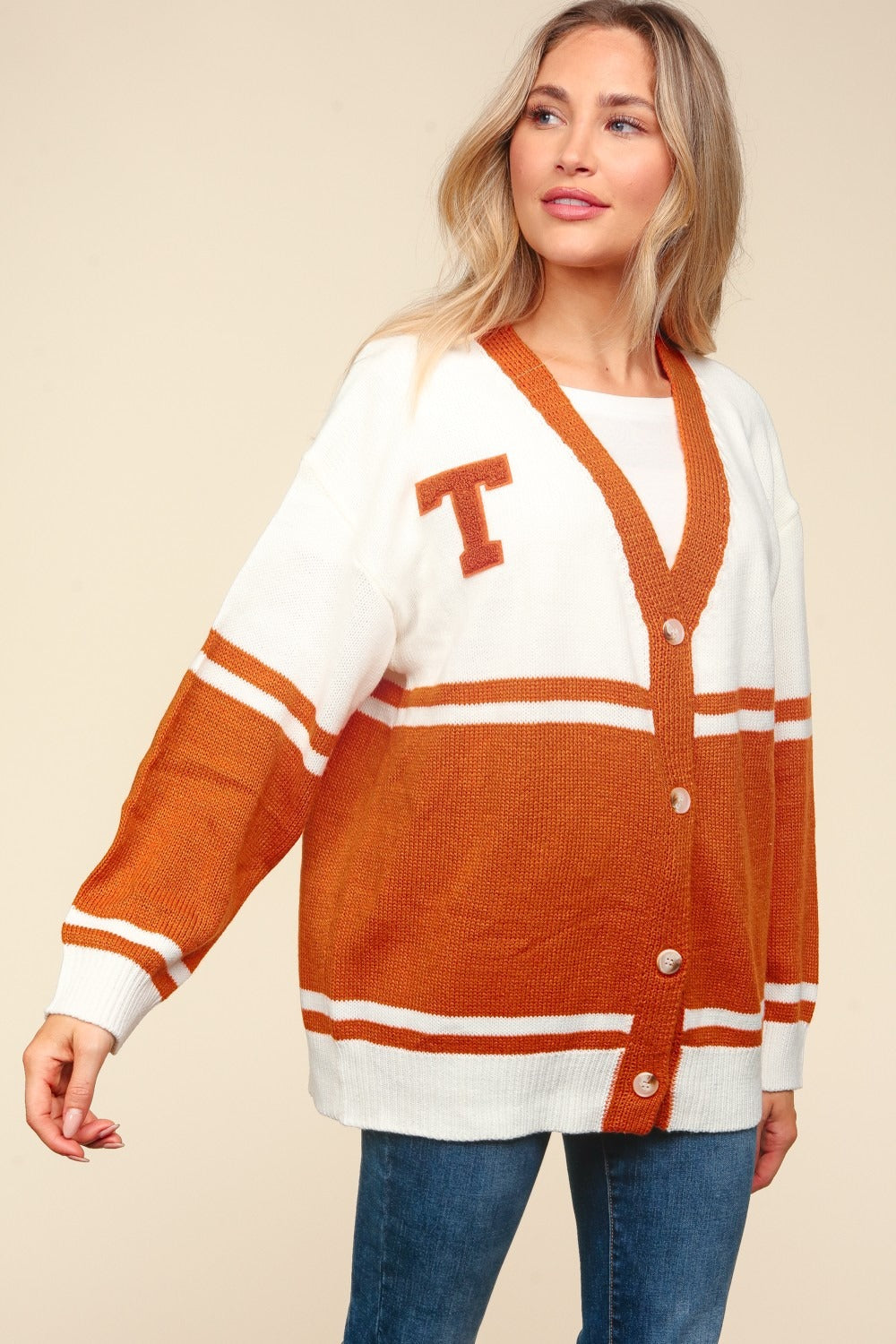 A person is dressed in the Haptics Full Size V Neck Button Down Letter Patch Cardigan, featuring an oversized design in cream and rust-orange colors with horizontal stripes and a prominent letter T patch on the front. The cardigan is buttoned down over a white top paired with jeans, creating an elegant look against a beige background.