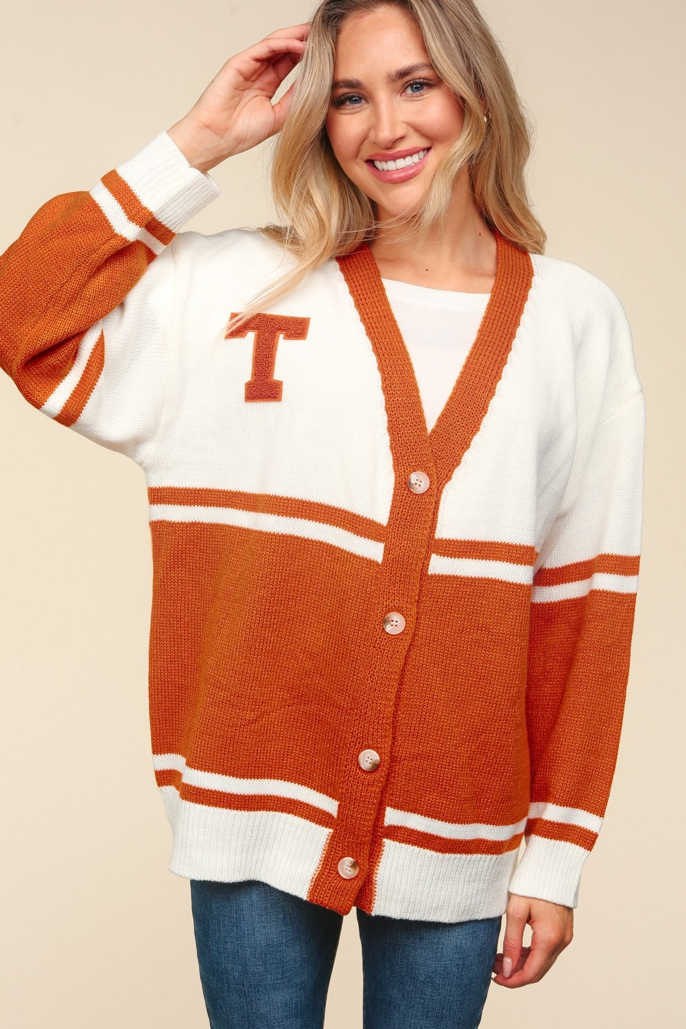 A person with long blonde hair smiles while wearing the Haptics Full Size V Neck Button Down Letter Patch Cardigan in white and orange, featuring a T patch on the chest over a white shirt. With one hand playfully touching their hair, they complete the look with oversized-fit blue jeans.