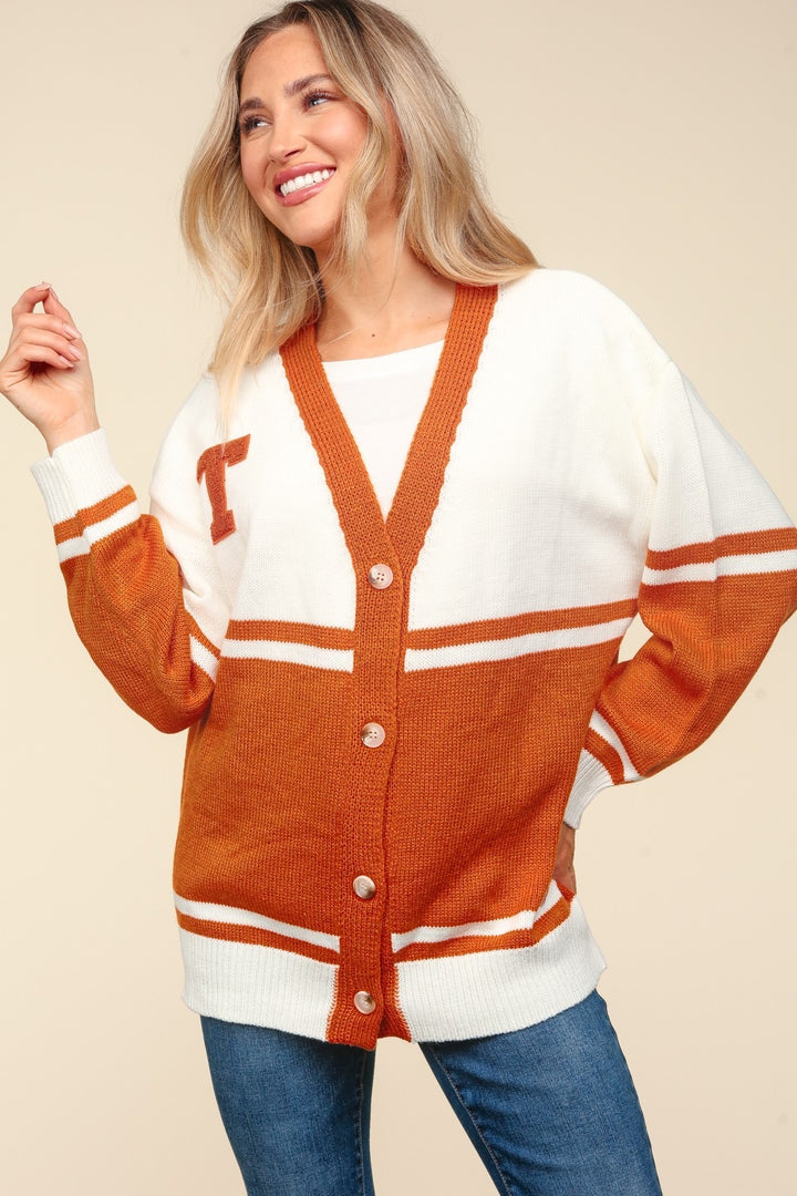 A smiling woman with long blonde hair wears the Haptics Full Size V Neck Button Down Letter Patch Cardigan in oversized white and orange. She pairs it with a white top underneath and blue jeans, set against a neutral beige background.