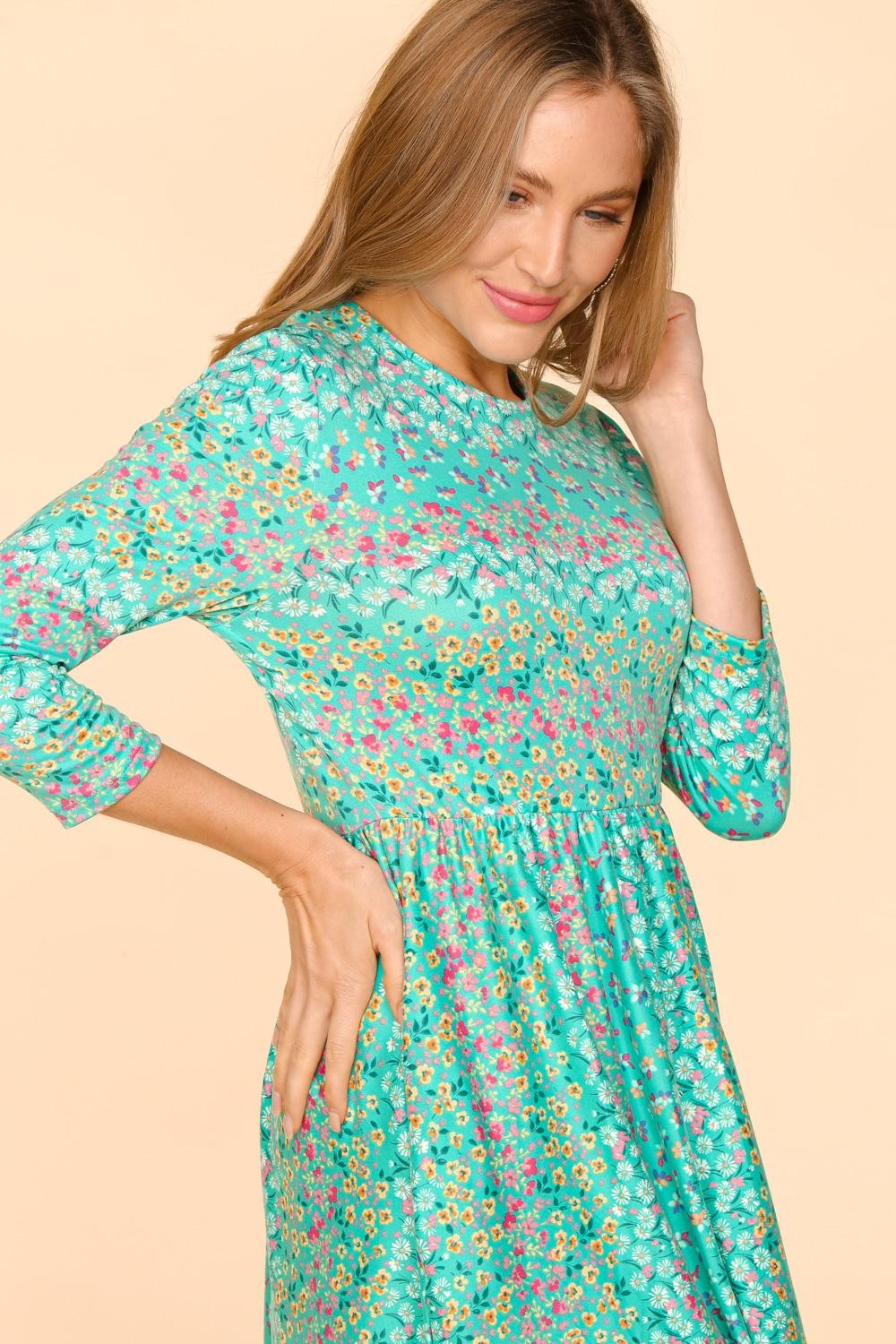 A woman with long hair wears a Haptics Round Neck Floral Dress with Pockets, featuring bright turquoise with pink, yellow, and white patterns. Its comfortable and breathable fabric ensures her ease as she poses gracefully against a plain beige background, smiling softly.