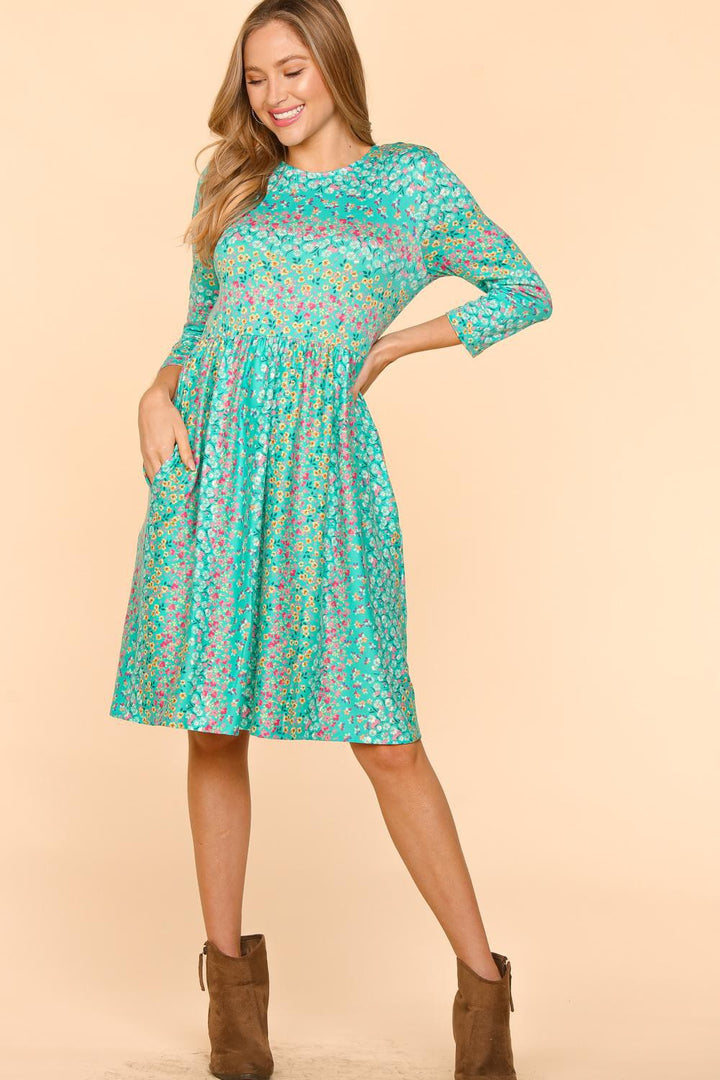 Wearing a comfortable Haptics Round Neck Floral Dress with Pockets, in teal floral print, three-quarter sleeves, and paired with brown ankle-high boots, a person poses against a beige backdrop with one hand on their hip and a smile.