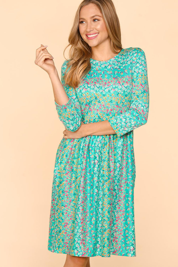 A woman smiles against a beige background in a teal Haptics Round Neck Floral Dress with handy pockets, 3/4 length sleeves, and a pleated skirt. Made from comfortable, breathable fabric, she holds a strand of her light brown hair in her right hand.