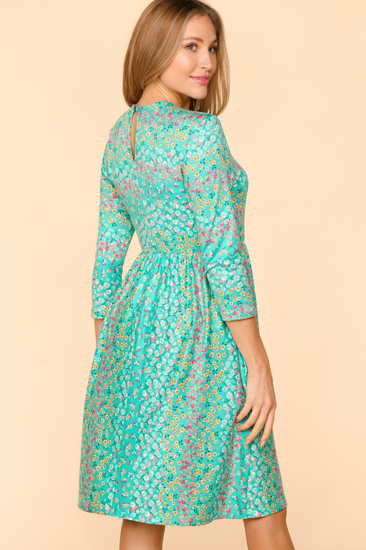 A woman in the Haptics Round Neck Floral Dress with three-quarter sleeves poses against a beige background, looking over her shoulder. Her light brown hair is styled down, highlighting the comfortable and breathable fabric of this knee-length outfit.