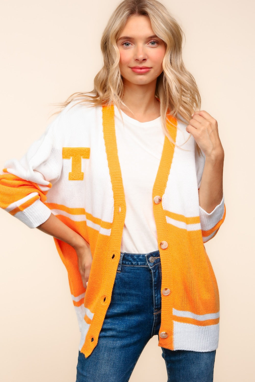 A woman with long blonde hair wears the Haptics V Neck Button Down Letter Patch Cardigan, which features a white and orange oversized design and a letter patch T. She pairs it with a white shirt and blue jeans, standing confidently with a soft smile while holding one side of the cardigan against a neutral background.