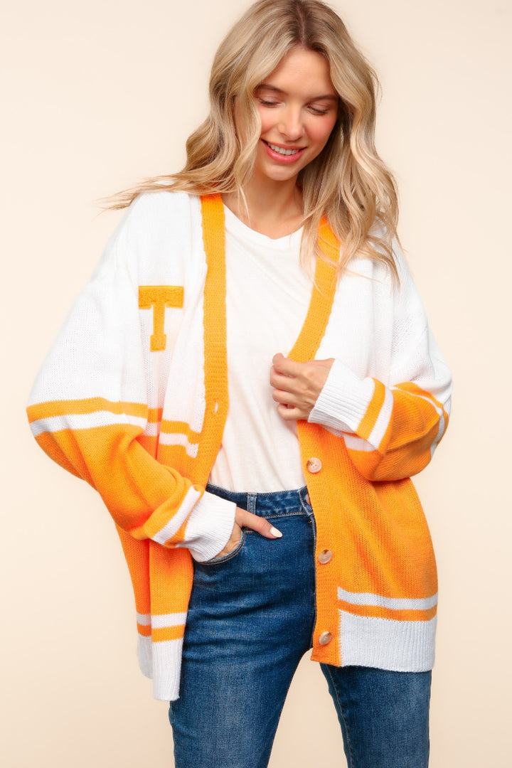 A smiling woman with long blonde hair wears the Haptics V Neck Button Down Letter Patch Cardigan, featuring a distinctive letter patch T on the chest, over a white shirt and blue jeans. She stands against a light background, with one hand in her pocket and the other resting casually on her stylish cardigan.