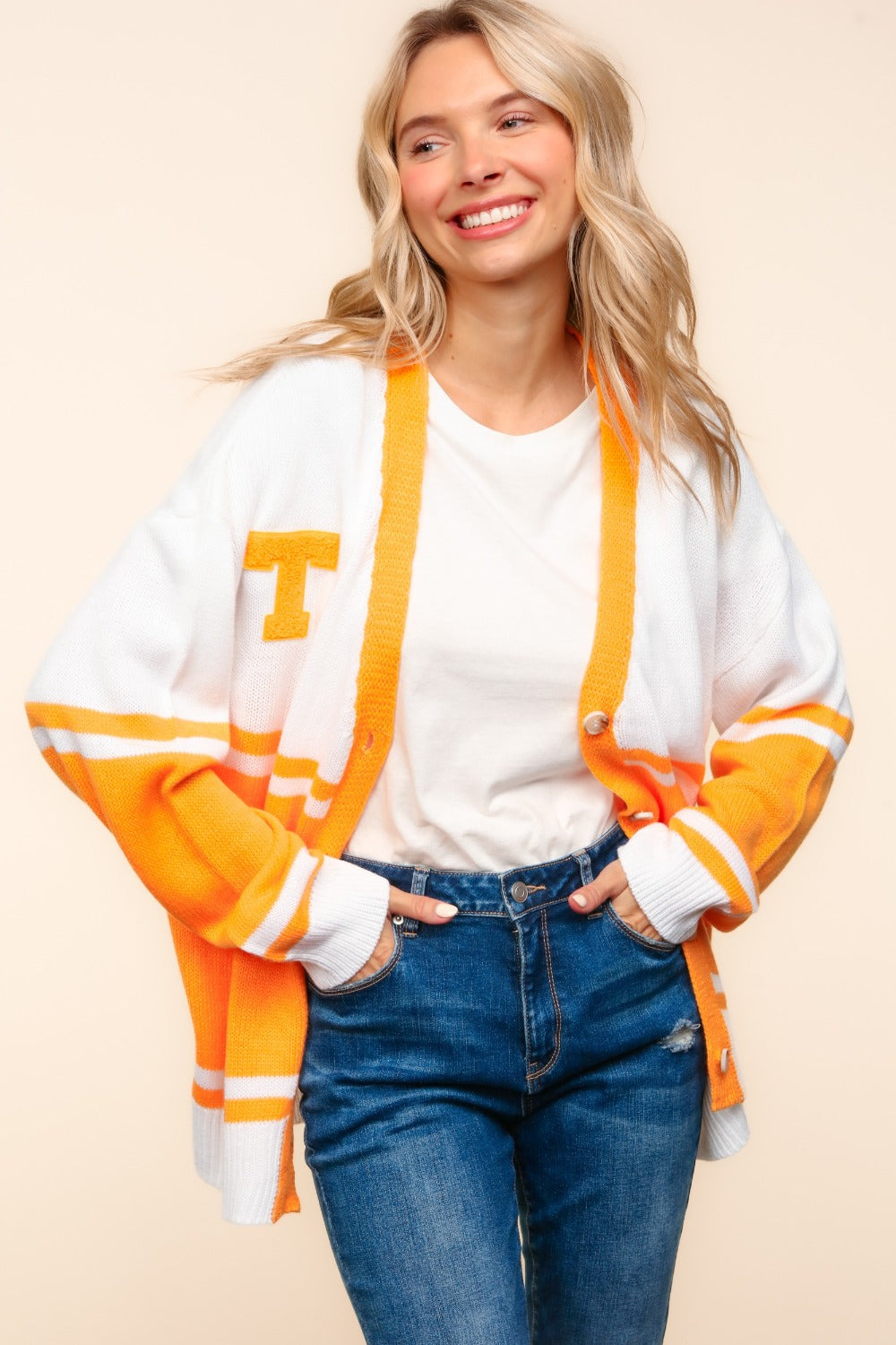 A person with long blonde hair smiles while wearing the Haptics V Neck Button Down Letter Patch Cardigan in white and orange, paired with a white shirt and blue jeans. Their hands are in their pockets as they stand against a plain beige background.