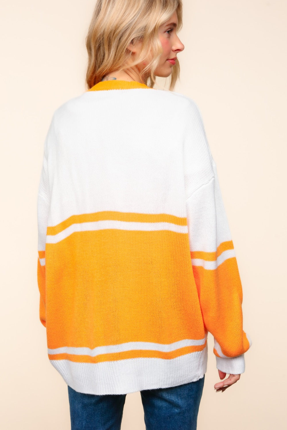 A woman wearing an oversized fit Haptics V Neck Button Down Letter Patch Cardigan, featuring white and orange stripes, stands against a neutral background. Her long blonde hair flows over her blue jeans, embodying a casual yet stylish vibe. The subtle letter patch adds a touch of uniqueness to this trendy look.