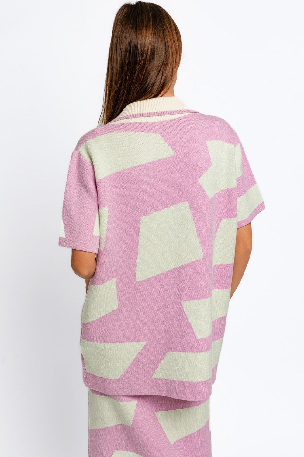 A person with long brown hair facing away wears the Le Lis Abstract Contrast Short Sleeve Collared Cardigan in a pink and white geometric pattern, paired with a matching skirt. The cardigan showcases a contrasting collar. The background is plain white.