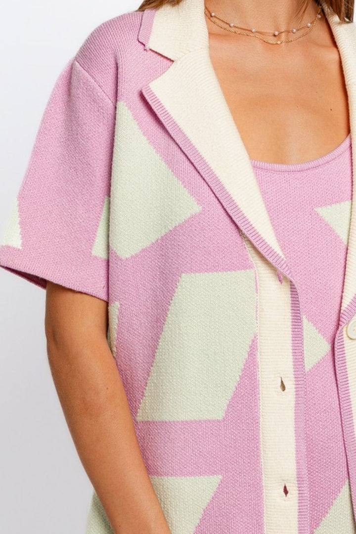 A woman models the Le Lis Abstract Contrast Short Sleeve Collared Cardigan, showcasing its light pink and cream geometric design over a coordinated top. The cardigan features short sleeves, cream lapels, and a partially buttoned front. The image focuses on the upper torso, without featuring the face.