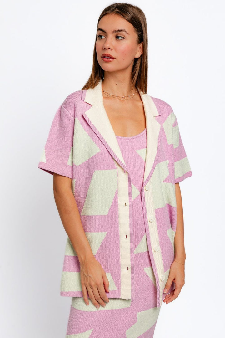 A woman is wearing the Le Lis Abstract Contrast Short Sleeve Collared Cardigan over a matching dress, both adorned with large geometric shapes in pink and cream. Her long hair sways gracefully as she looks to the side against a plain white background.