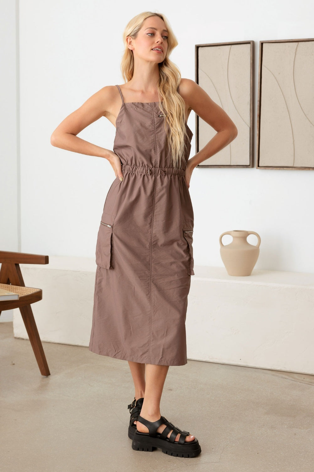 A woman with long blonde hair is wearing a utilitarian chic Le Lis Adjustable Strap Waist Drawstring Cargo Midi Dress in brown, paired with black platform sandals, as she stands in a minimalist room. Abstract paintings and a ceramic vase adorn the shelf in the background.