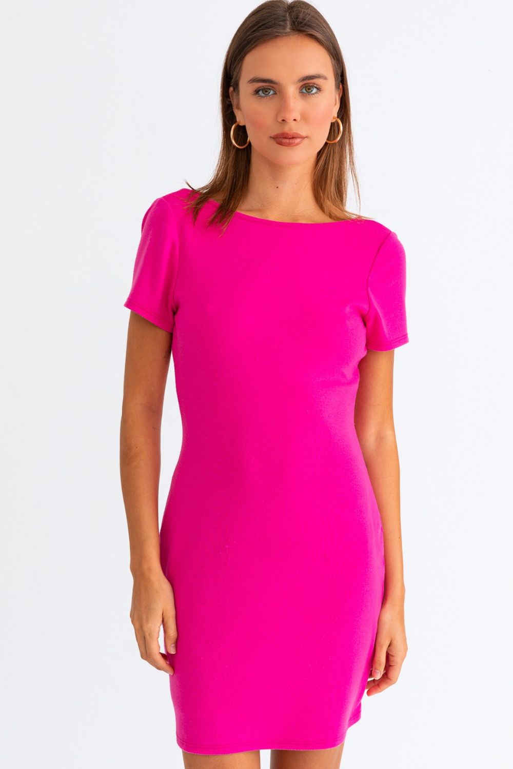 A woman stands against a plain white background, wearing the Le Lis Short Sleeve Low Back Mini Dress in bright pink. Her long brown hair cascades down her back, framed by hoop earrings, as she looks directly at the camera.