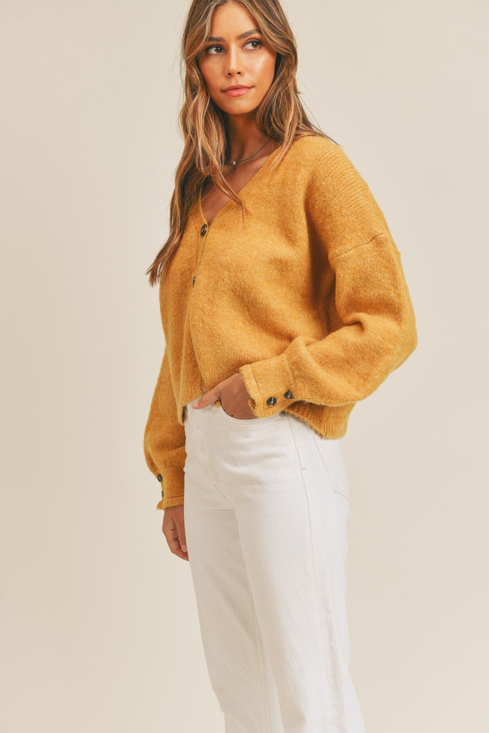 A woman with long hair wears the MABLE Long Sleeve Button Down Sweater Cardigan in yellow, paired with white pants, as she stands against a plain background. Her right hand is in her pocket and she looks to the side.