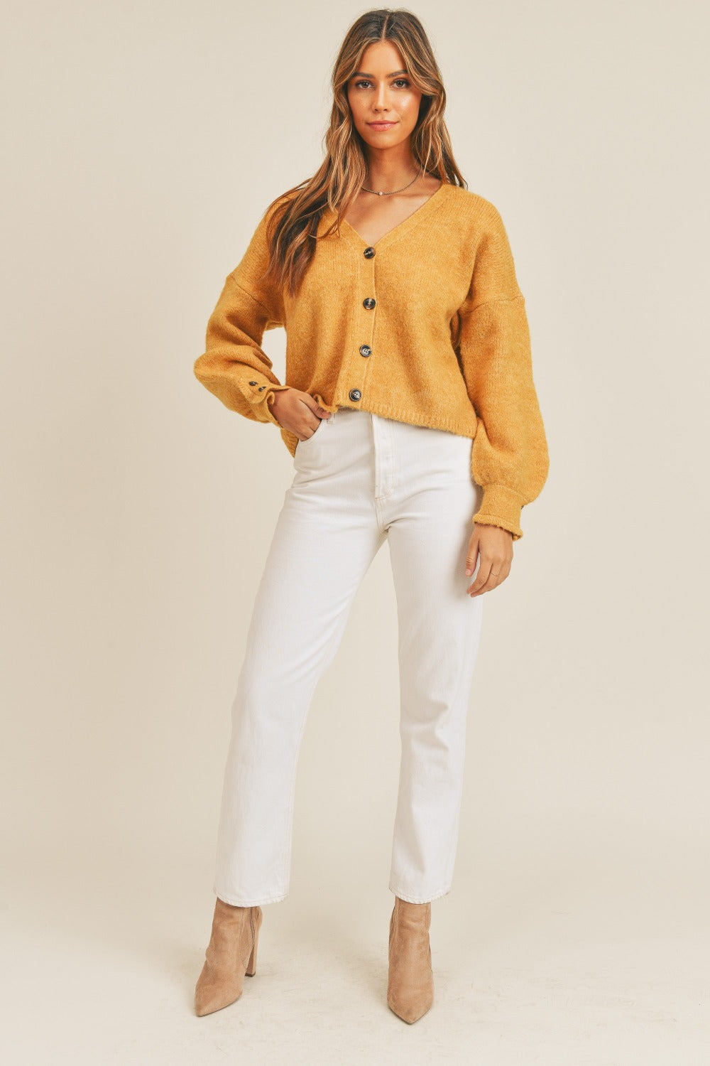 A person wearing the MABLE Long Sleeve Button Down Sweater Cardigan in a mustard color, paired with white jeans and beige ankle boots, stands against a plain background. They have long hair and are looking at the camera with a slight smile.