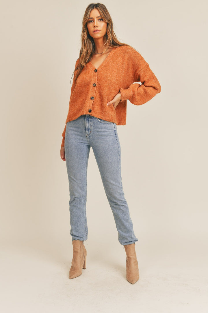 A woman stands against a plain background wearing the MABLE Long Sleeve Button Down Sweater Cardigan in a rust color, paired with light blue jeans and tan ankle boots. Her long hair falls in loose waves, and she exudes confidence with one hand casually tucked into her pocket.