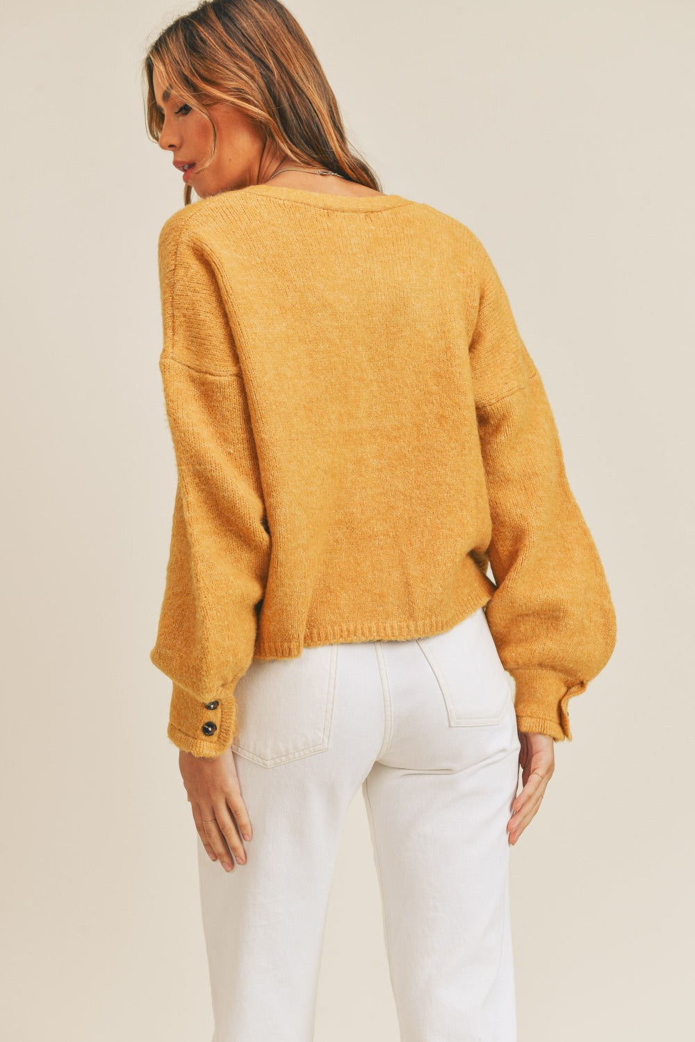 A person with long hair is wearing the mustard yellow MABLE Long Sleeve Button Down Sweater Cardigan, crafted from knit fabric, and cream-colored pants. The cardigan features balloon sleeves with button details on the cuffs. The person is turned slightly to the side against a plain background.