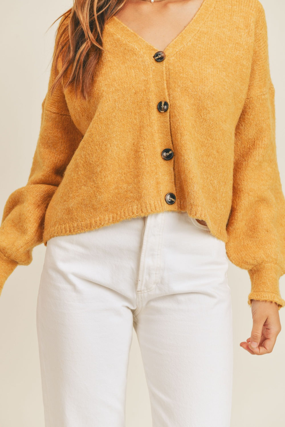 A person is wearing the MABLE Long Sleeve Button Down Sweater Cardigan in mustard yellow, crafted from knit fabric, paired with white pants. The cardigan features large black buttons and a slightly loose fit. The person's face is not visible in the image.