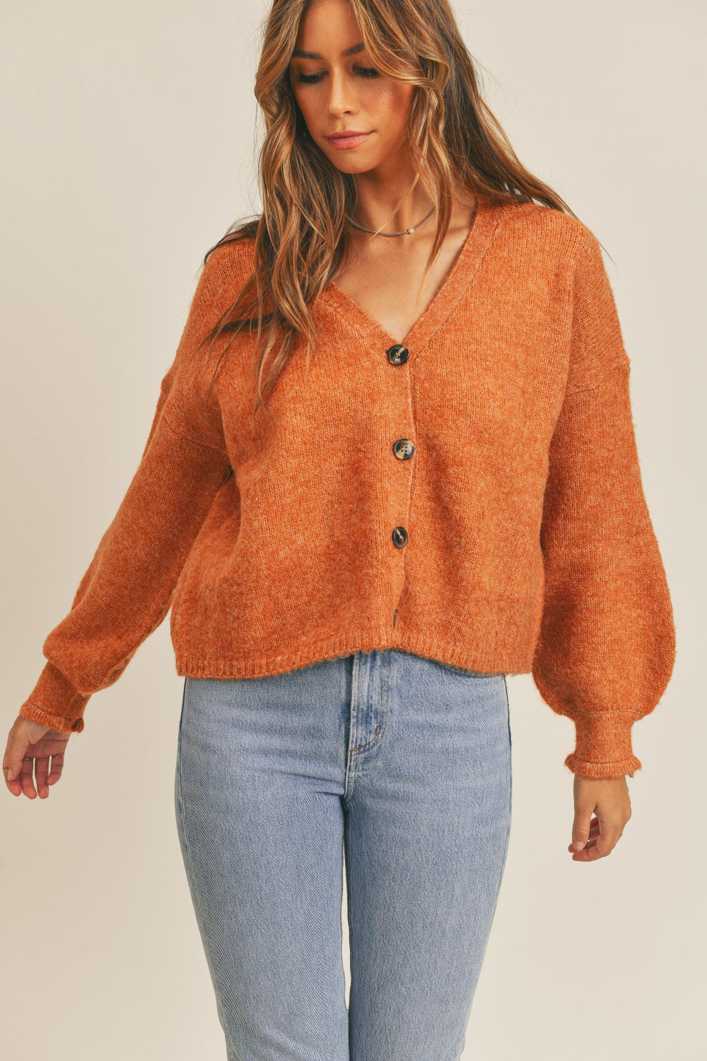 A woman with long hair is pictured wearing the MABLE Long Sleeve Button Down Sweater Cardigan, which is crafted from textured orange knit fabric, paired with light blue jeans. The V-neck cardigan, featuring stylish balloon sleeves, stands out against a neutral background as she gazes slightly to the side.