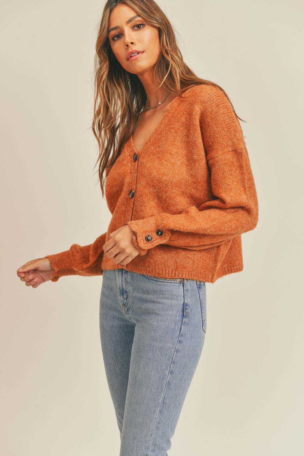 A person wearing the MABLE Long Sleeve Button Down Sweater Cardigan in a rust hue, combined with light blue jeans, stands against a plain background. They have long, wavy hair and are looking at the camera.