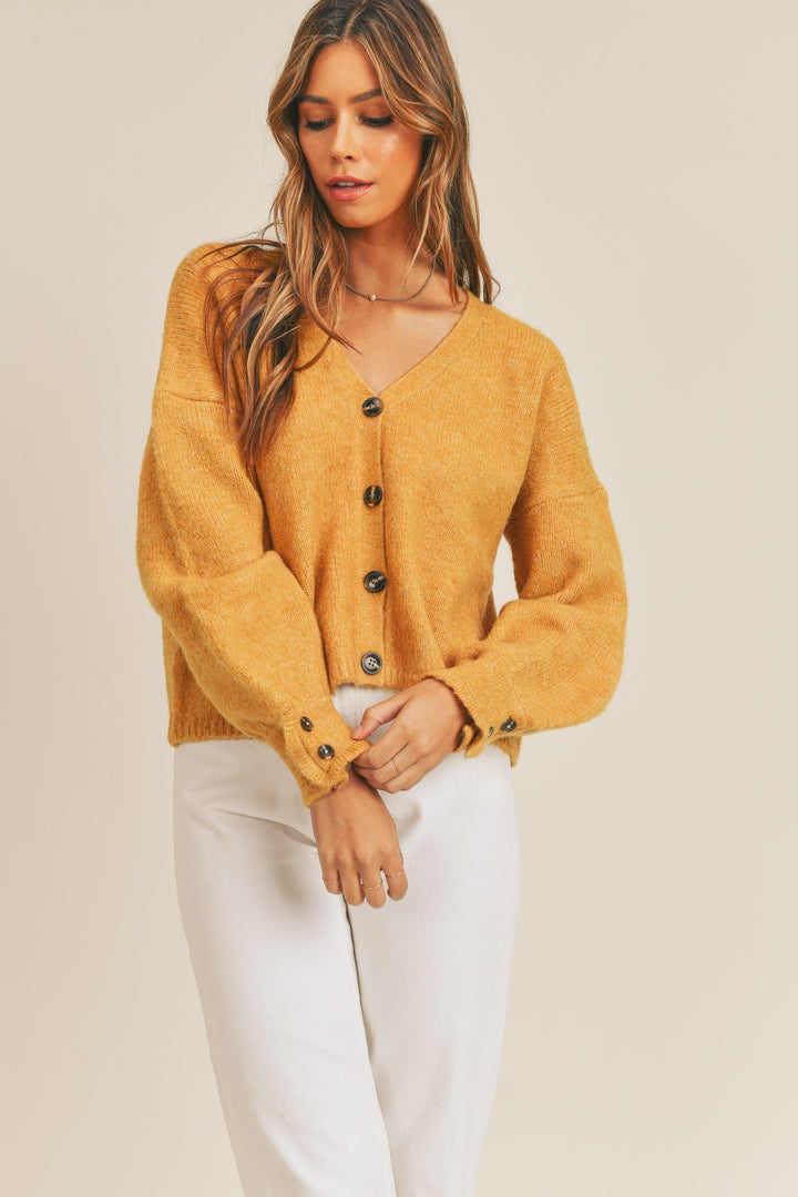 A person wearing the MABLE Long Sleeve Button Down Sweater Cardigan in mustard yellow, made from knit fabric and featuring black buttons, pairs it with white pants as they stand against a neutral background. Their long hair falls as they look slightly downward, with one hand gently touching the cardigan.