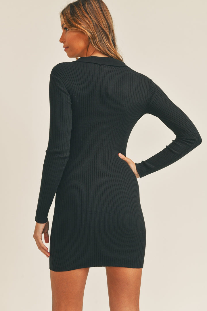 A person is wearing the fitted black Mable Button Up Ribbed Mini Shirt Dress, featuring long sleeves and a high neck, facing away. The dress falls above the knee and is set against a plain off-white background, emphasizing its versatility and blend of elegance with simplicity.