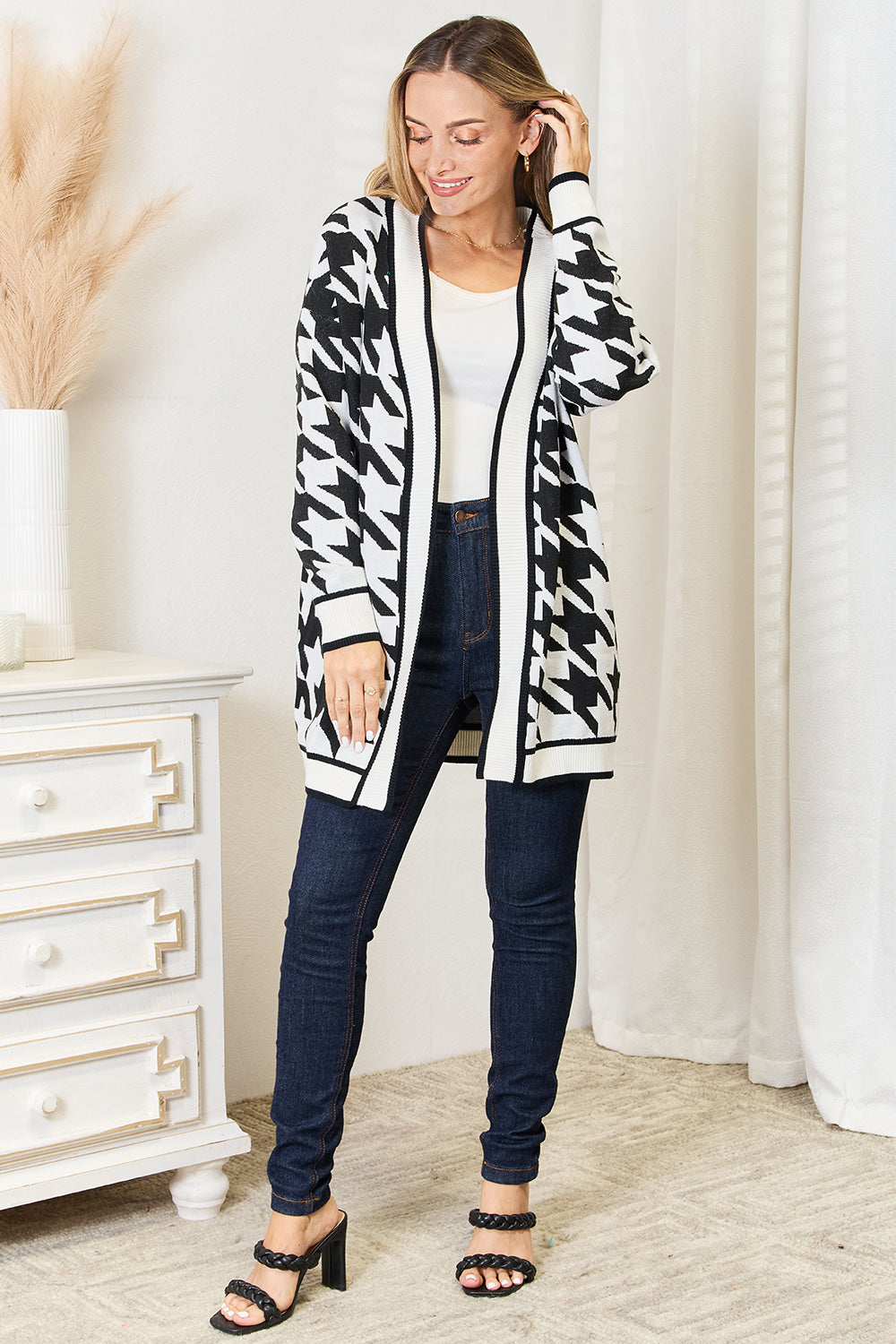 A person smiling and looking down is wearing a Mandy Woven Right Houndstooth Open Front Longline Cardigan over a white top, paired with dark jeans and black sandals. They stand near a white dresser with a vase of pampas grass.