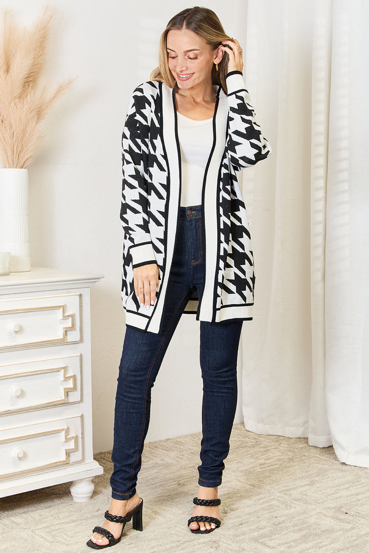 A person smiling and looking down is wearing a Mandy Woven Right Houndstooth Open Front Longline Cardigan over a white top, paired with dark jeans and black sandals. They stand near a white dresser with a vase of pampas grass.