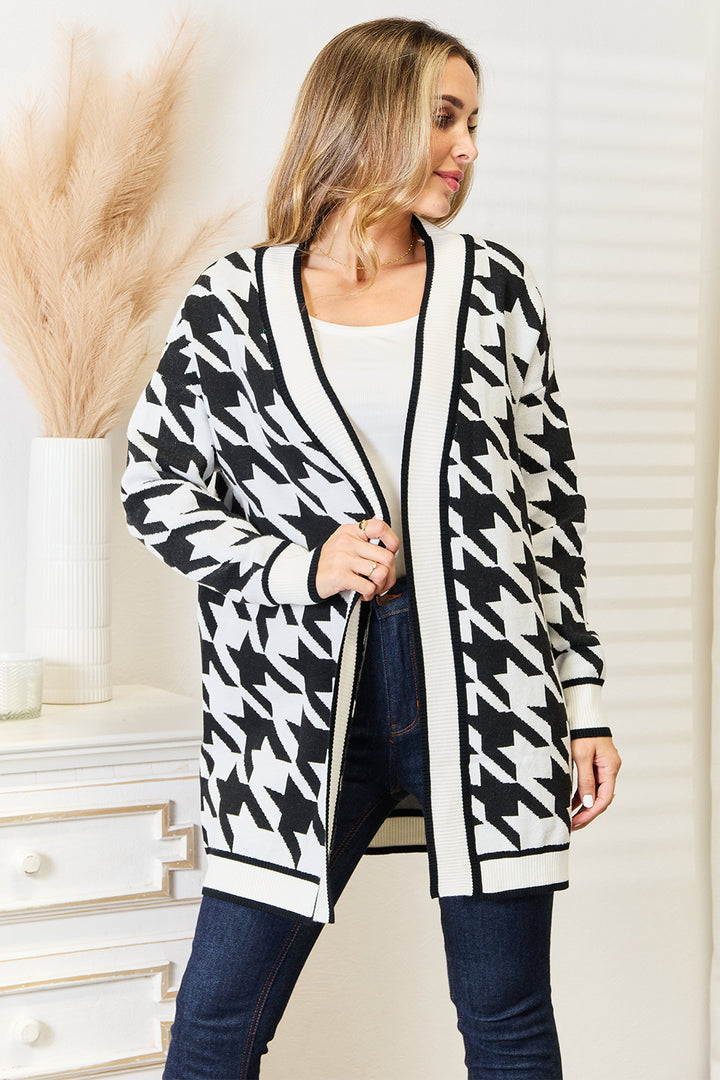 A woman confidently poses in the softly lit room, wearing the Mandy Woven Right Houndstooth Open Front Longline Cardigan over a white top and dark jeans. With one hand on her hip, she stands near a white dresser adorned with decorative beige pampas grass.