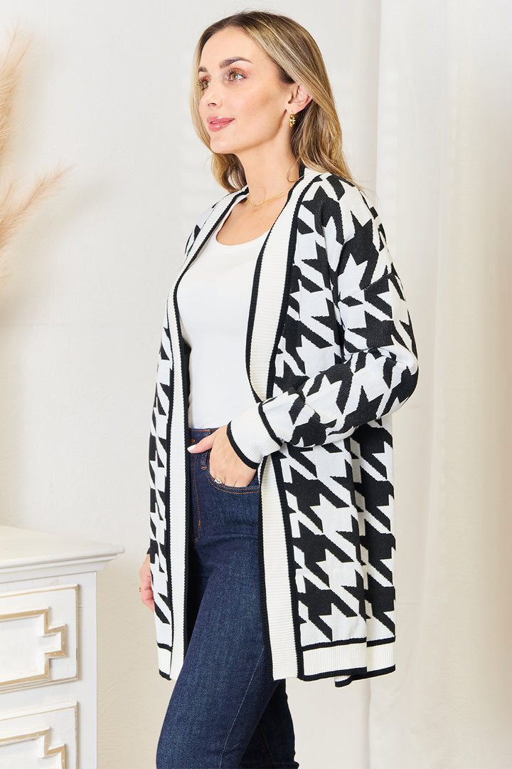 A person dressed in the Mandy Woven Right Houndstooth Open Front Longline Cardigan over a white top and blue jeans is standing indoors. They are positioned in front of a light-colored wall with a decorated table nearby.