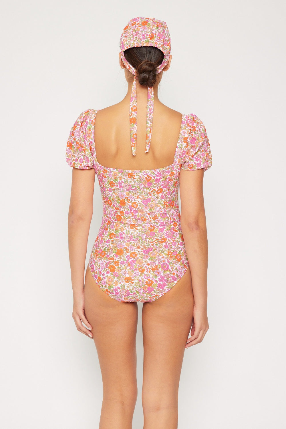 Wearing the Marina West Swim Floral Puff Sleeve One-Piece, a person faces away, showcasing the elegant square neckline and puff sleeves. The suit is adorned with pink and orange flowers on a light background, complemented by a matching cap tied at the back.