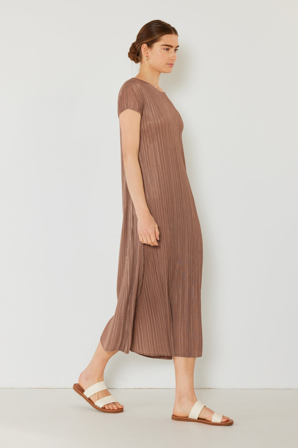 Wearing the Marina West Swim Pleated Cap Sleeve A-Line Dress and sandals, a woman elegantly showcases her feminine charm against a simple backdrop.