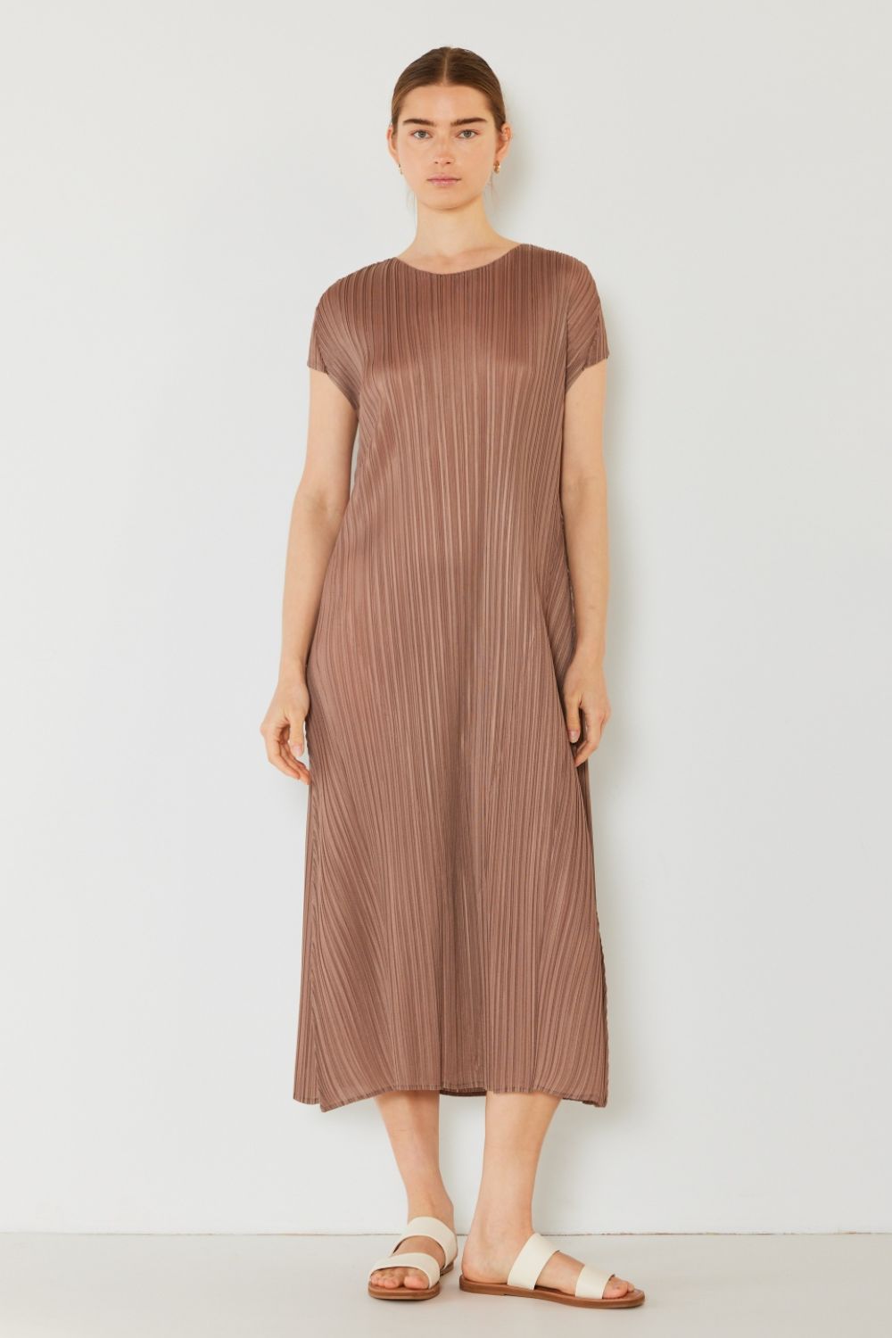 A person stands gracefully against a white background in the Marina West Swim Pleated Cap Sleeve A-Line Dress, exuding feminine charm. With long, dark hair tied back and light-colored open-toed sandals, they have their arms relaxed by their sides.