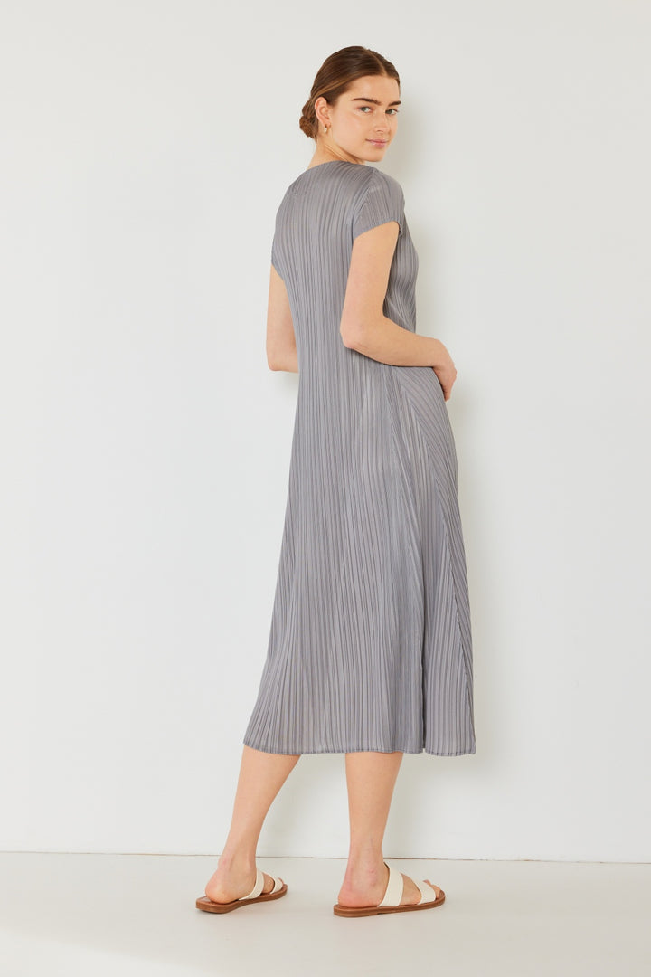 A woman in a Marina West Swim Pleated Cap Sleeve A-Line Dress stands against a white wall, exuding feminine charm. She turns slightly to the side, looks over her shoulder, and wears white sandals.