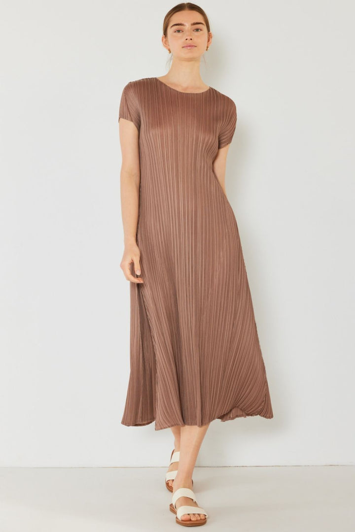 Against a white backdrop, someone in a Marina West Swim Pleated Cap Sleeve A-Line Dress and white sandals stands with hair pulled back, gazing forward. The brown dresss feminine charm is highlighted by its mid-calf length and cap sleeves.