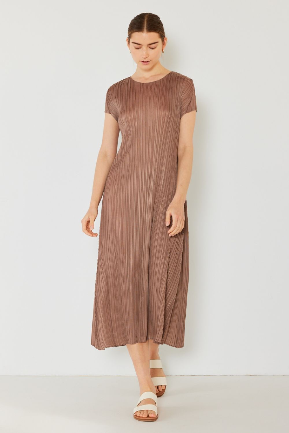 Dressed in a Marina West Swim Pleated Cap Sleeve A-Line Dress, radiating feminine charm with its brown pleats, they pair it elegantly with white sandals against a plain, light-colored background.